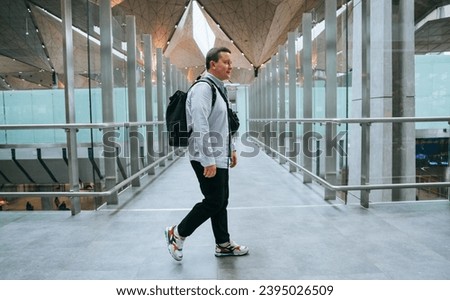 Male traveler with backpack walking through airport terminal. Young Caucasian man in casual clothes goes to flight gate. Travel, journey, trip, relocation. Active blogger at modern building. Royalty-Free Stock Photo #2395026509