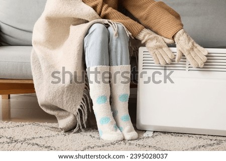 Young woman in winter clothes with warm plaid near electric heater at home. Concept of heating season Royalty-Free Stock Photo #2395028037