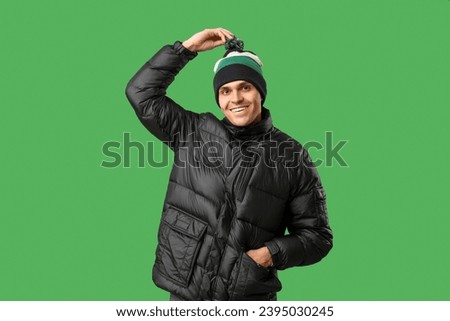 Young man in stylish puffer jacket and hat on green background