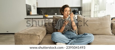 Portrait of stylish young woman on sofa, eating cereals, holding bowl and spoon, watching tv and having a quick breakfast, spending time at home in living room. Royalty-Free Stock Photo #2395041493
