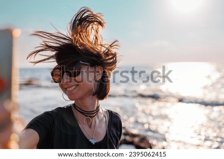 Close-up of a stunning woman with hair blowing in the wind taking a selfie by the sea. Beautiful girl smiling while looking at camera outside - fashionable lifestyle and beauty concept