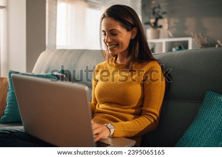 A cheerful woman is sitting on sofa at her comfortable apartment with a laptop in her lap and scrolling on the phone. A young brunette is using technologies for online chat and hanging on social media Royalty-Free Stock Photo #2395065165