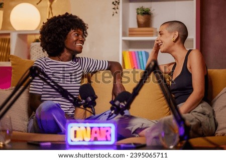Two diverse women recording a podcast while using a microphone and sitting on a cozy sofa in a modern room. Female podcaster making audio podcast from her home studio and interviewing her guest.