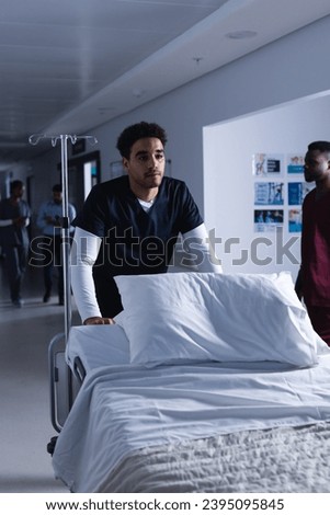 Biracial male doctor walking with bed in hospital corridor. Medicine, healthcare and medical services, unaltered.