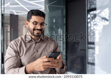 Businessman holding phone inside office, joyful man smiling uses smartphone app at workplace, browses social networks, and writes text message.