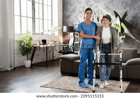 Happy pensioner grandmother elderly patient with walking frame with assistance of nurse caregiver at home. Walking after trauma injury Royalty-Free Stock Photo #2395115155