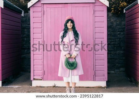 Stylish young smiling hipster woman with color hair wearing pink coat, knitted hat and fur bag on pink wooden beach hut background. Seasonal city street fashion. Barbiecore style. Selective focus. Royalty-Free Stock Photo #2395122817