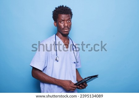 Male healthcare professional with stethoscope stands in profile, holding a digital tablet. African American nurse wears scrubs and carries modern technology to address COVID-19 concerns.