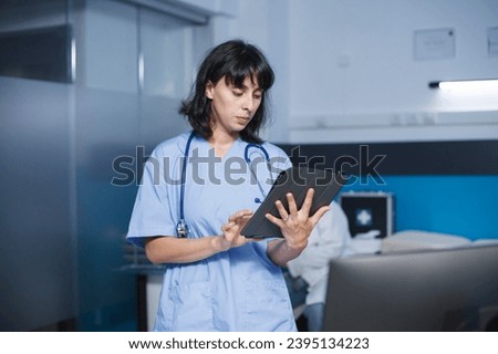 Detailed image of a caucasian woman in blue scrubs standing in a clinic office using a tablet for medical research. Female nurse utilizing a digital device to review patient appointments.