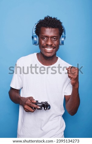 Portrait of excited black male gamer with wireless headset, smiling and playing video games on console. Joyful African American man in casual clothing, celebrating victory while holding a controller.