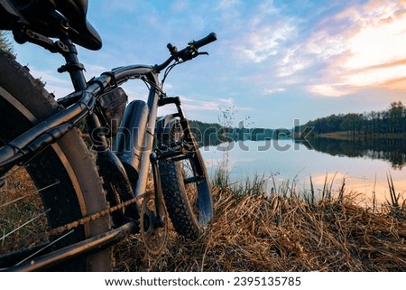 Electric bike with thick wheels on the background of lake and sunset. Sports fatbike. Picturesque place in the village. The concept of a healthy lifestyle.