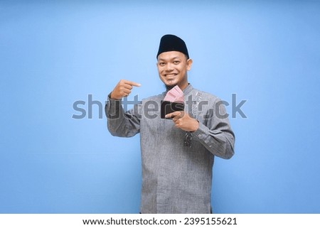 Asian Muslim man holding wallet of full cash money ready to pay zakat isolated on blue background. Islamic lifestyle concept Royalty-Free Stock Photo #2395155621