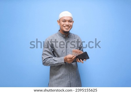 Asian Muslim man holding wallet of full cash money ready to pay zakat isolated on blue background. Islamic lifestyle concept Royalty-Free Stock Photo #2395155625