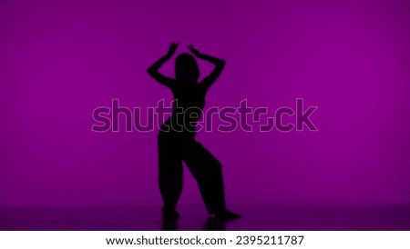 In the frame on the purple background in the silhouette. Dances slender, beautiful girl. Demonstrates dance moves in the style of hip hop, lifting hands up. It is feminine, plastic, rhythmic