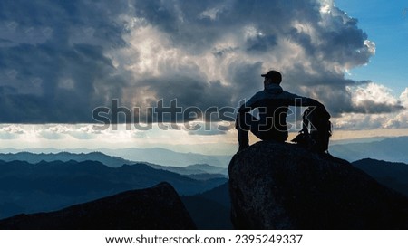 Low angle view of Asian male hikers climbing mountain at sunset rays over the clouds with trekking poles sitting on cliff edge on top of rock mountain, Successful summit concept image.   Royalty-Free Stock Photo #2395249337