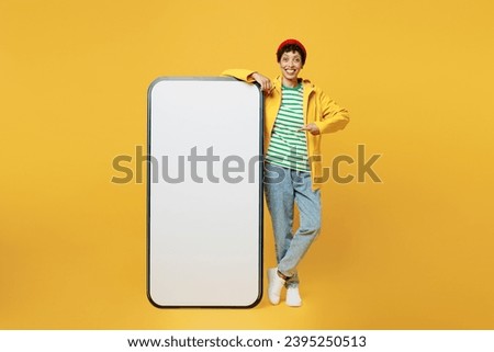Full body young woman wears raincoat outerwear red hat point finger on big huge blank screen mobile cell phone isolated on plain yellow background. Outdoors lifestyle wet fall weather season concept
