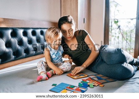 Mom teaches a little girl to collect puzzles in a cardboard tablet while sitting on the bed