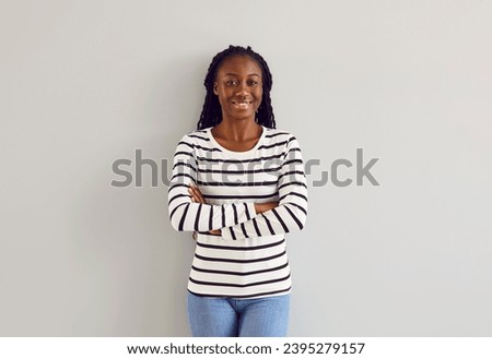 Portrait of beautiful female model in casual clothes. Happy young African American woman wearing white black striped long sleeve shirt and blue jeans standing with folded arms by light gray wall Royalty-Free Stock Photo #2395279157