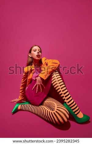 Portrait of stylish young girl in orange stripped tights, jacket and heel shoes, posing against magenta color studio background. Concept of retro fashion, queer, surrealism, art, beauty, ad. Royalty-Free Stock Photo #2395282345