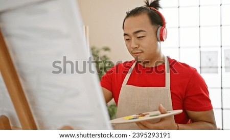 Serious young chinese man with pigtail hairstyle, artist standing in university art studio, effortlessly mastering the art of drawing while intently listening to music on headphones.