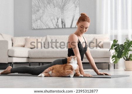 Beautiful woman with cute red cat practicing yoga on mat at home Royalty-Free Stock Photo #2395312877