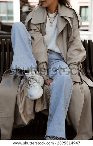 beautiful blonde girl dressed in beige oversized trench coat, blue palazzo jeans, white sweater, massive sneakers and bag, details of stylish fashion outfit, lifestyle model sitting Royalty-Free Stock Photo #2395314947