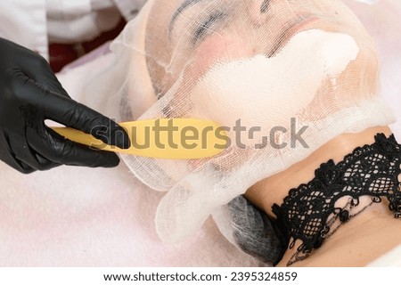 A beautician uses a spatula to apply cream to an already prepared face with gauze, a procedure for facial skin with couperosis. Royalty-Free Stock Photo #2395324859