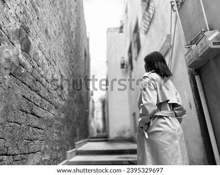 Black and white photo of a young girl in an autumn coat walks the street lonely in an old city part. Old bricks walls, stairs in the narrow street. Street walker. Beautiful girl alone is going.