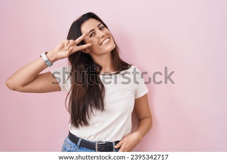Young brunette woman standing over pink background doing peace symbol with fingers over face, smiling cheerful showing victory 