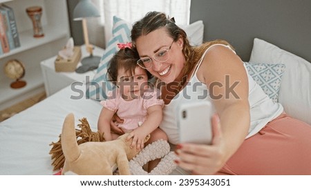 Mother and daughter love, sitting on the comfy bed in their room, smiling, taking a relaxed morning selfie picture with a smartphone