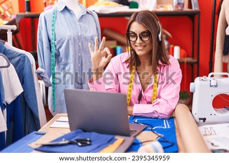 Young hispanic woman dressmaker designer doing video call with laptop doing ok sign with fingers, smiling friendly gesturing excellent symbol 
