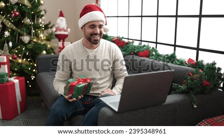 Young hispanic man having video call holding gift sitting by christmas tree at home