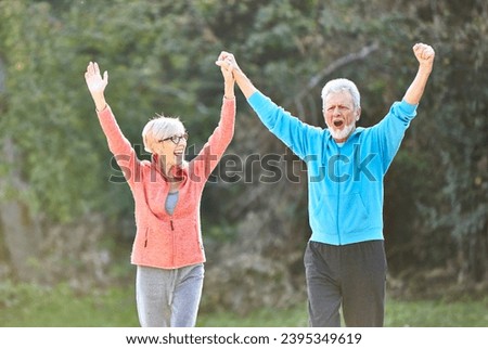 Smiling active senior couple jogging exercising and having fun and celebrating success rasing hands together taking a break in the park Royalty-Free Stock Photo #2395349619