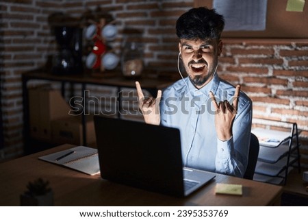 Young hispanic man with beard working at the office at night shouting with crazy expression doing rock symbol with hands up. music star. heavy concept. 