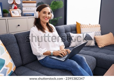 Young beautiful hispanic woman using laptop and headphones sitting on sofa at home
