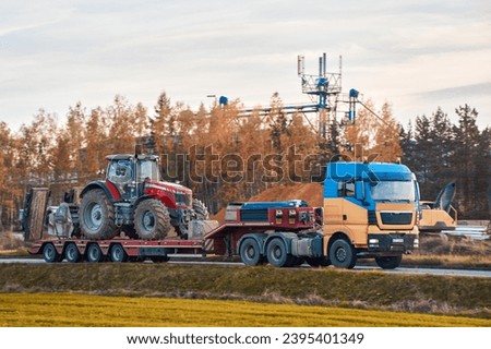 The semi truck is hauling farm equipment on a flatbed. The heavy industrial truck semi trailer flatbed platform transport one big modern farming tractor machine on the road. Evening sun. Royalty-Free Stock Photo #2395401349