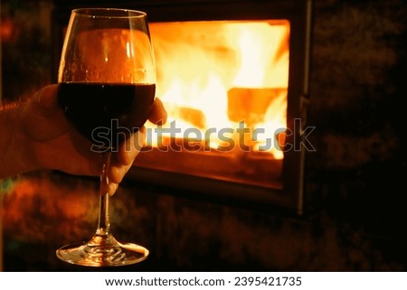 Mans hand holds the wine glass in front of the fireplace. Man with glass of wine near fireplace at home. Blur burning fireplace background.