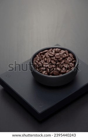 Dark roasted coffee beans a small black ceramic bowl on a black digital scale.