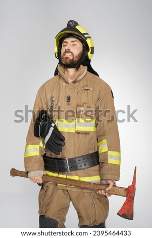 Exhausted caucasian firefighter with closed eyes, holding shabby axe. Front view of tired, bearded man in uniform with hatchet resting, on gray studio background. Concept of job, workplace, equipment.