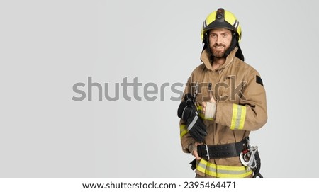 Cheerful male firefighter in uniform gesturing with thumb up. Front view of smiling bearded fireman giving like, while looking at camera, isolated on gray, copy space. Concept of work, hand gesture. Royalty-Free Stock Photo #2395464471