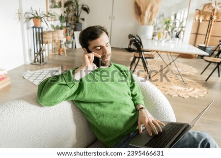 Bearded freelancer in green jumper and jeans talking on smartphone while using laptop at home