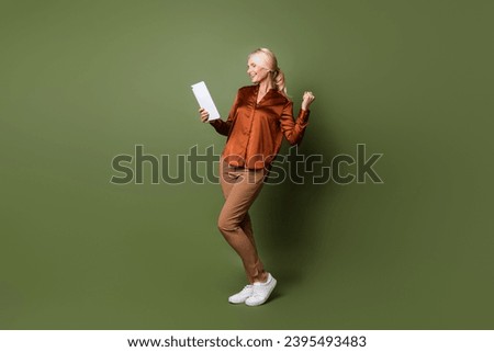 Full body photo of attractive pensioner woman hold tablet fist celebrate wear trendy brown satin clothes isolated on khaki color background