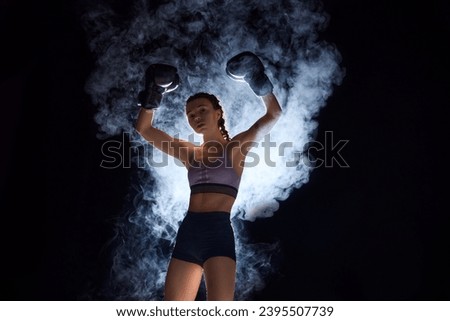 Powerful boxer girl in shorts and gloves vining pose, symbolizing strength and competitiveness in sports against black background in stage smoke. Sport, active lifestyle, health, competition concept.