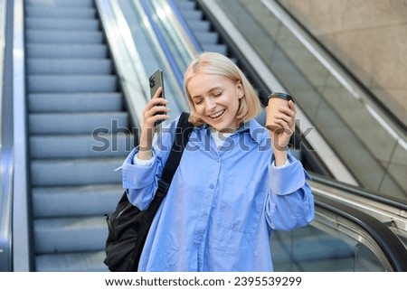 Image of young happy blond woman, dancing from happiness and joy, drinking takeaway coffee, holding smartphone, posing near escalator in city centre. Royalty-Free Stock Photo #2395539299