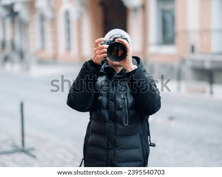 Male photographer takes a photo using professional digital camera against background of city street. Royalty-Free Stock Photo #2395540703