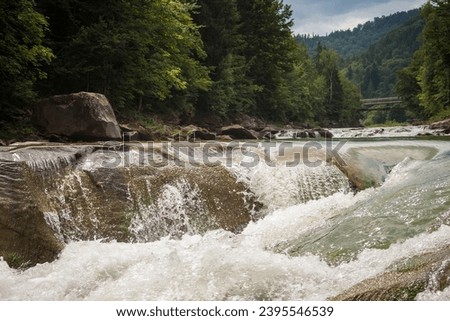 Mountain river with a waterfall. The rapid flow of water flows between the wooded shores