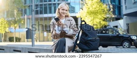 Lifestyle portrait of cute blonde girl with smartphone, sitting on bench with backpack, using mobile phone, reading message.