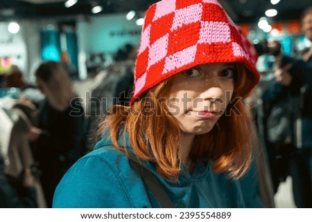 Close-up portrait of an adorable red-haired teenage girl wearing a funny red knitted hat, with a joyful expression, creating a cute and playful atmosphere during the winter season. Royalty-Free Stock Photo #2395554889