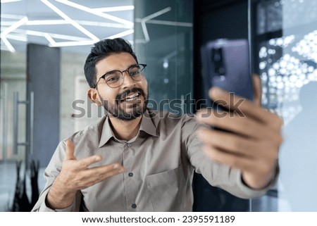 Young successful businessman at workplace inside office talking on video call using app on phone, close up man at workplace smiling.