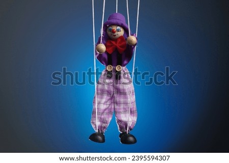 The frame features a puppet in a blue background, a marionette that hangs from strings. Someone is holding it and controlling it. Depicts a theatrical performance, a clown who smiles